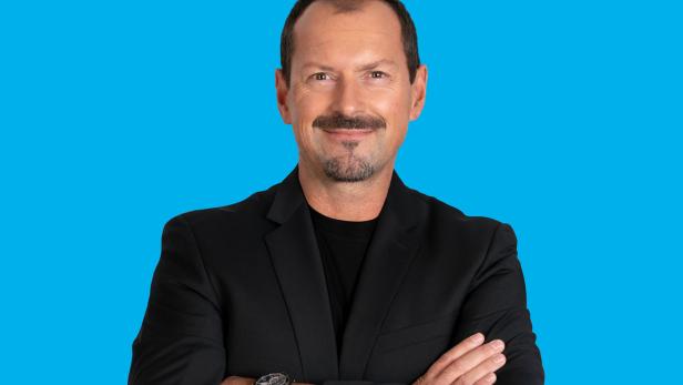 Ein Mann mit Schnurrbart und Anzug vor einem blauen Hintergrund.