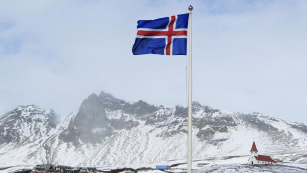 Die isländische Flagge weht vor einer schneebedeckten Landschaft mit Bergen und einer kleinen Kirche.