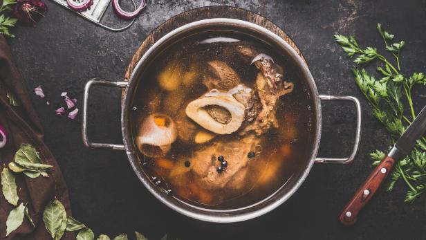 Ein Topf mit Knochenbrühe, Zwiebeln und Kräutern auf einem dunklen Untergrund.