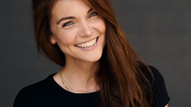 Eine lächelnde Frau mit braunem Haar und einem schwarzen T-Shirt vor einem grauen Hintergrund.