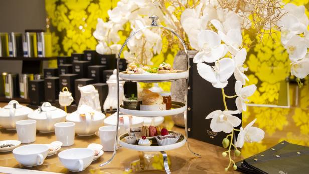 Ein dreistöckiger Servierständer mit verschiedenen Köstlichkeiten vor einem Hintergrund aus Blumen und Teegeschirr.