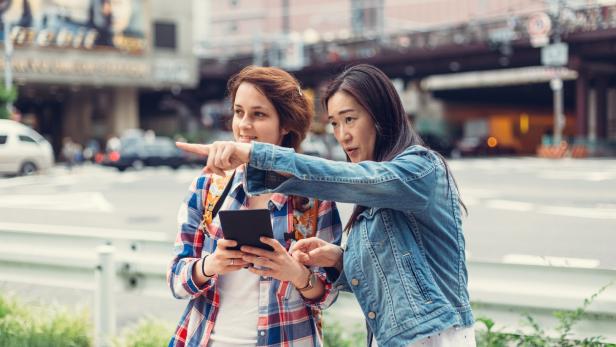 Zwei Frauen stehen in einer Stadt und schauen auf ein Tablet, während eine von ihnen in die Ferne zeigt.