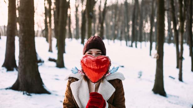 Eine Frau mit roter Mütze hält einen herzförmigen Ballon vor dem Gesicht in einem verschneiten Wald.
