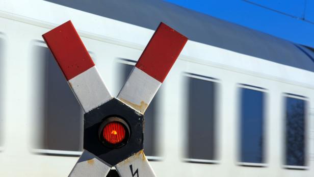 Ein Andreaskreuz mit rotem Blinklicht vor einem vorbeifahrenden Zug.