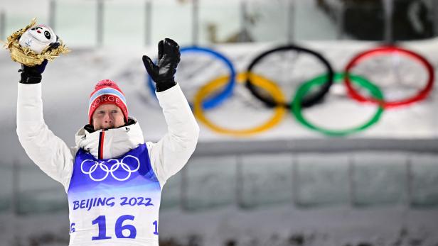 Ein Biathlet feiert mit der Bing Dwen Dwen-Maskottchen-Trophäe vor den Olympischen Ringen in Peking 2022.