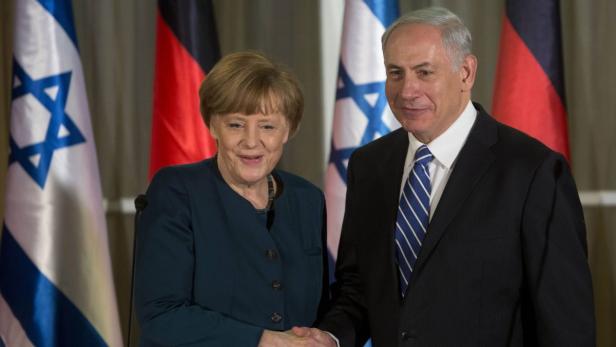 Angela Merkel und Benjamin Netanjahu schütteln sich vor israelischen und deutschen Flaggen die Hände.