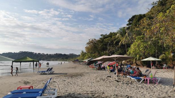 Ein Strand mit Liegestühlen, Sonnenschirmen und Menschen unter bewölktem Himmel.
