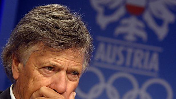 Ein Mann mit nachdenklichem Gesichtsausdruck vor einem Hintergrund mit dem Schriftzug „Austria“ und den olympischen Ringen.