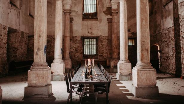 Ein langer Tisch mit Gedeck steht in einem Raum mit Säulen und Ziegelwänden.