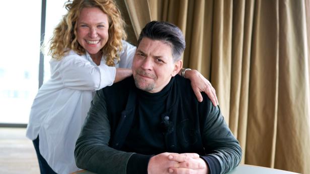 Eine lächelnde Frau legt ihre Hand auf die Schulter eines Mannes, der an einem Tisch sitzt.