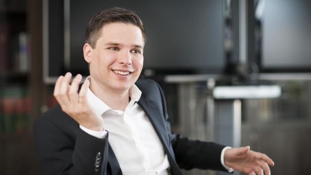 Ein lächelnder Mann im Anzug gestikuliert vor einem unscharfen Hintergrund.