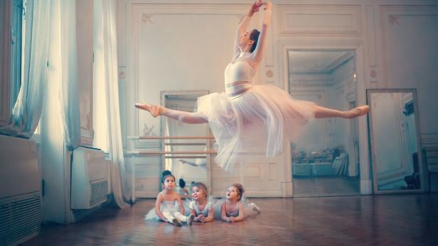 Eine Ballerina springt vor drei kleinen Mädchen in einem Ballettsaal.