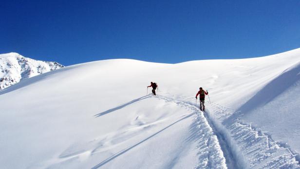 Zwei Skibergsteiger steigen einen verschneiten Hang unter blauem Himmel hinauf.