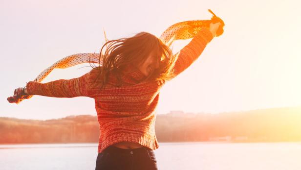 Eine Frau mit Schal steht mit ausgestreckten Armen vor einem See.