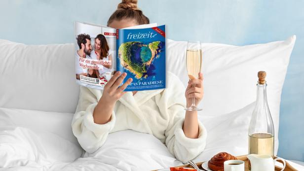 Eine Frau liest im Bett eine Zeitschrift und hält ein Glas Sekt in der Hand.