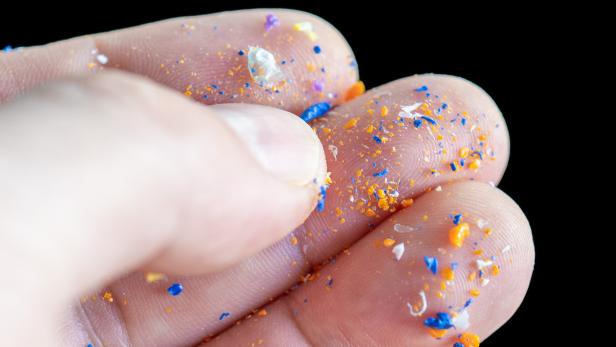 Eine Hand hält kleine bunte Plastikteilchen, hauptsächlich in Blau, Orange und Weiß, auf den Fingerspitzen.