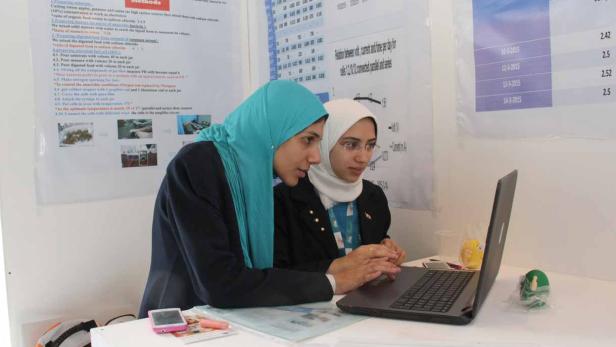 Zwei junge Frauen mit Kopftüchern arbeiten an einem Laptop vor einem wissenschaftlichen Poster.