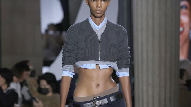 Eine Frau auf dem Laufsteg präsentiert ein Outfit mit bauchfreiem Oberteil und Faltenrock.