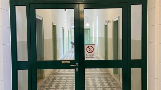 Eine grüne Doppeltür mit Glas führt in einen Flur mit Schachbrettmuster und einem "Rauchen verboten"-Schild.