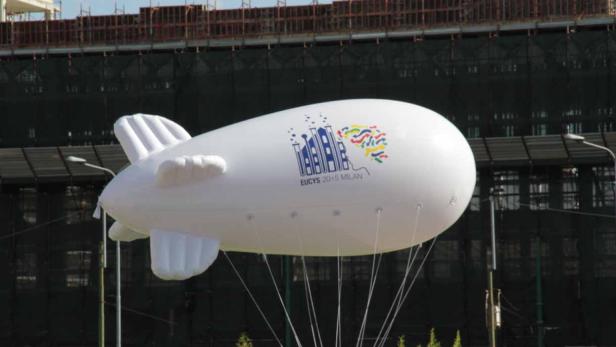 Ein weißer Werbe-Zeppelin mit dem Logo von EUCYS 2015 Mailand vor einem Gebäude.