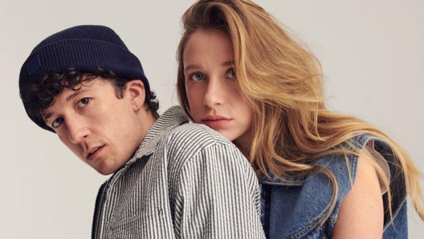 Ein Mann mit Mütze und eine Frau in Jeanskleidung posieren vor einem hellen Hintergrund.