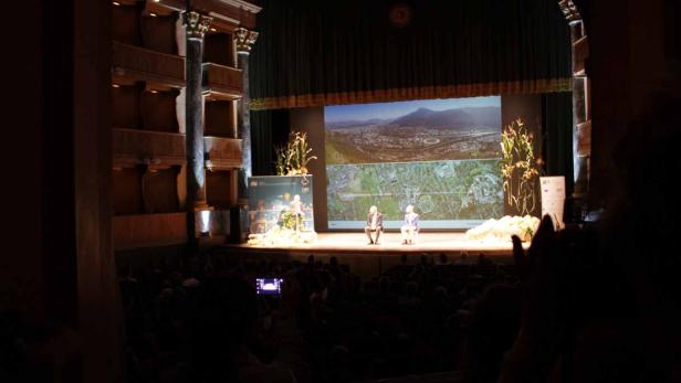 Zwei Personen sitzen auf einer Bühne vor einem großen Publikum.
