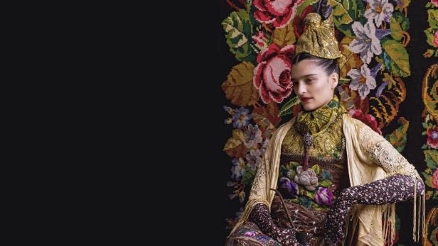 Eine Frau in traditioneller Tracht sitzt vor einem blumigen Wandteppich.