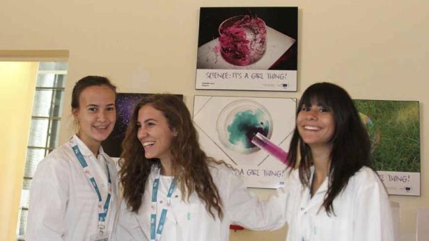 Drei junge Frauen in Laborkitteln lächeln vor Postern mit wissenschaftlichen Motiven.