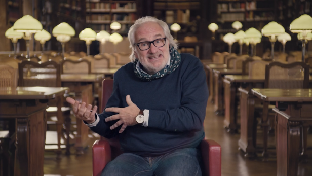 Ein Mann mit Brille gestikuliert in einer Bibliothek sitzend.