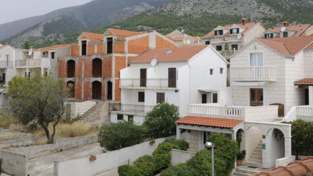 Blick auf weiße Wohnhäuser mit roten Ziegeldächern vor einem bewaldeten Hügel.