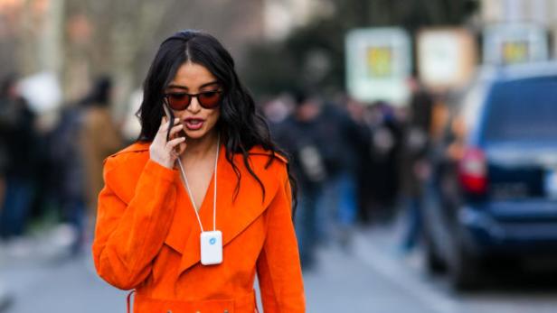 Eine Frau in einem orangefarbenen Mantel und passenden Schuhen telefoniert auf der Straße.