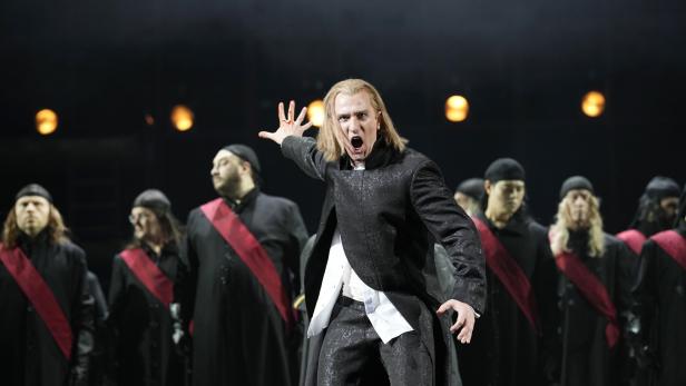 Ein Mann in einem schwarzen Anzug schreit auf einer Bühne, umgeben von Chormitgliedern.