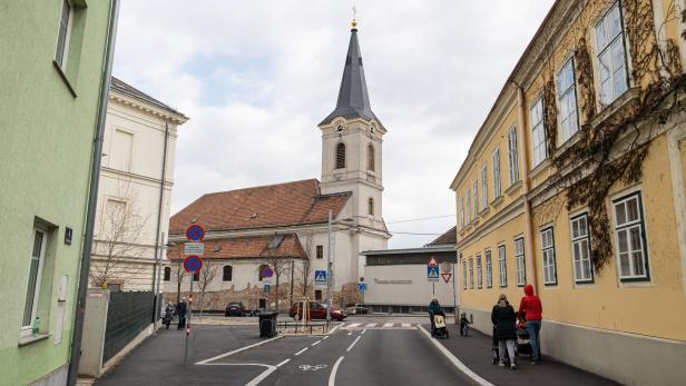 Eine Kirche mit hohem Turm steht an einer Straße, umgeben von alten Gebäuden und mehreren Fußgängern.