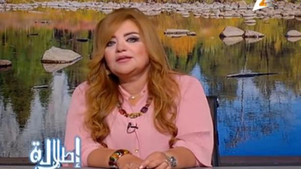 Eine Frau mit blonden Haaren sitzt vor einem Hintergrund mit einem See und Bäumen.