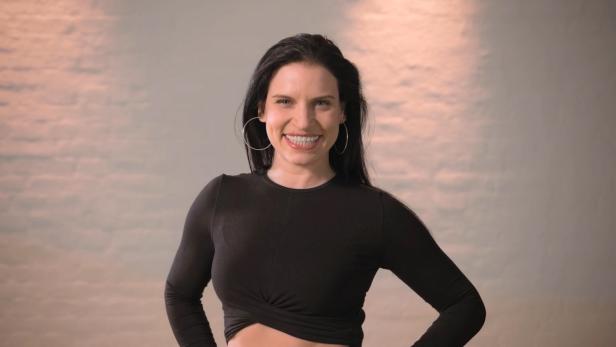 Eine lächelnde Frau mit langen, dunklen Haaren und schwarzen Oberteil posiert vor einem hellen Hintergrund.