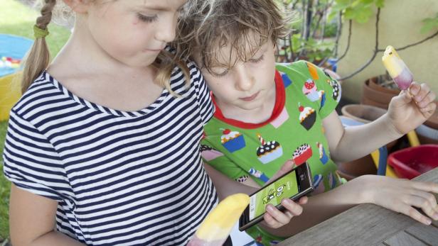 Zwei Kinder betrachten ein Smartphone und halten Eis am Stiel in der Hand.