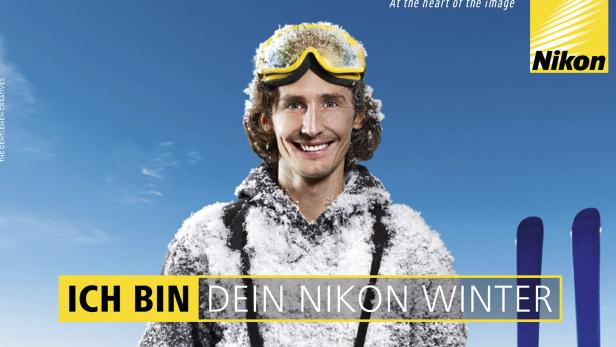 Ein lächelnder Mann mit Skibrille und Schnee auf der Kleidung vor blauem Himmel.