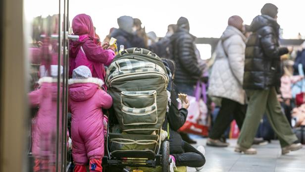 Zwei Kinder in Winterkleidung stehen neben einem Kinderwagen in einer belebten Fußgängerzone.