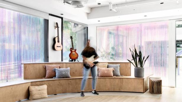 Eine unscharfe Person spielt Gitarre in einem Raum mit Gitarren an der Wand und bunten Vorhängen.