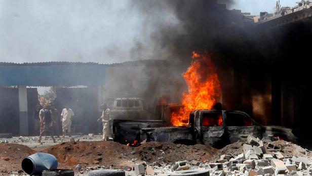 Ein brennendes Auto steht vor einem beschädigten Gebäude, Rauch steigt auf.