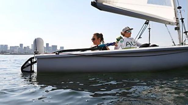 Zwei Seglerinnen in einem Segelboot auf dem Wasser vor einer Stadtkulisse.