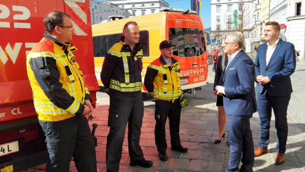 Eine Gruppe von Feuerwehrleuten und Anzugträgern steht vor Feuerwehrfahrzeugen.