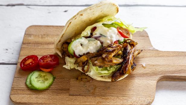 Ein Pita-Sandwich mit Fleisch, Salat, Tomaten, Gurken und Soße auf einem Holzbrett.