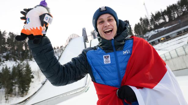 Ein lächelnder Skispringer mit österreichischer Flagge und Maskottchen vor einer Skisprungschanze.