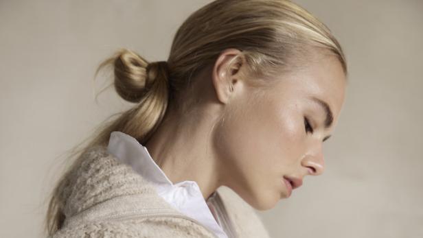 Eine blonde Frau trägt eine weiße Bluse unter einer beigen Strickweste.