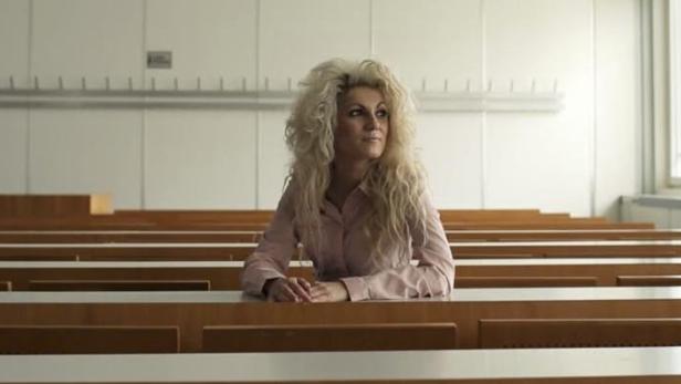 Eine Frau mit blonden Locken sitzt in einem leeren Hörsaal.
