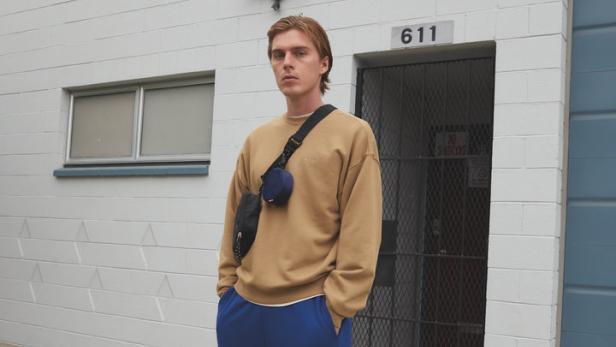 Ein Mann in beiger Kleidung und blauen Jogginghosen steht vor einem Gebäude.