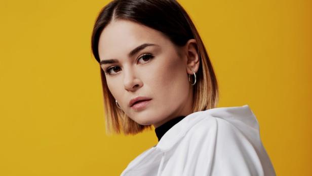 Eine Frau mit kurzem, braunem Haar und weißer Jacke vor einem gelben Hintergrund.