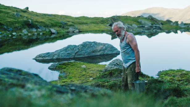Ein älterer Mann mit Bart trägt einen Eimer an einem Bergsee.
