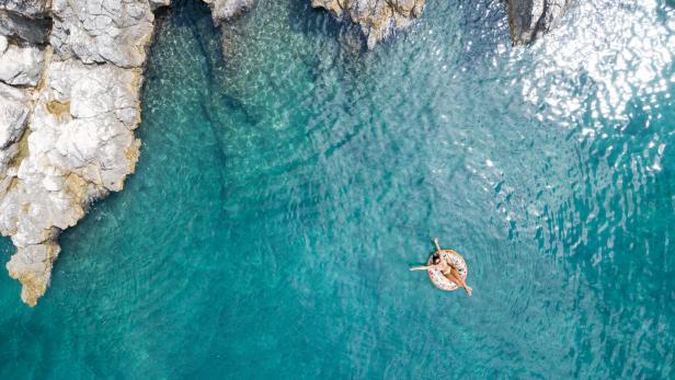 Eine Frau entspannt sich auf einer Luftmatratze im türkisfarbenen Wasser nahe einer felsigen Küste.